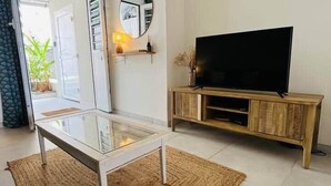 Ein Wohnzimmer mit Glastisch, Fernseher auf Holzständer, Spiegel an der Wand und einer Tür zum Balkon.