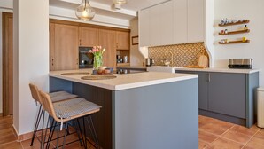 A kitchen with wooden cabinets, a central island, bar stools, pendant lighting, and a patterned backsplash.