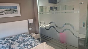 Una camera d'albergo con un letto, un comodino, un box doccia in vetro e un'opera d'arte a tema spiaggia.