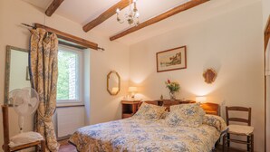 Ein Schlafzimmer mit einem Bett, zwei Nachttischen, einem Sessel, einem Ventilator, einem Fenster mit Vorhängen und einer Chandeliebre.
