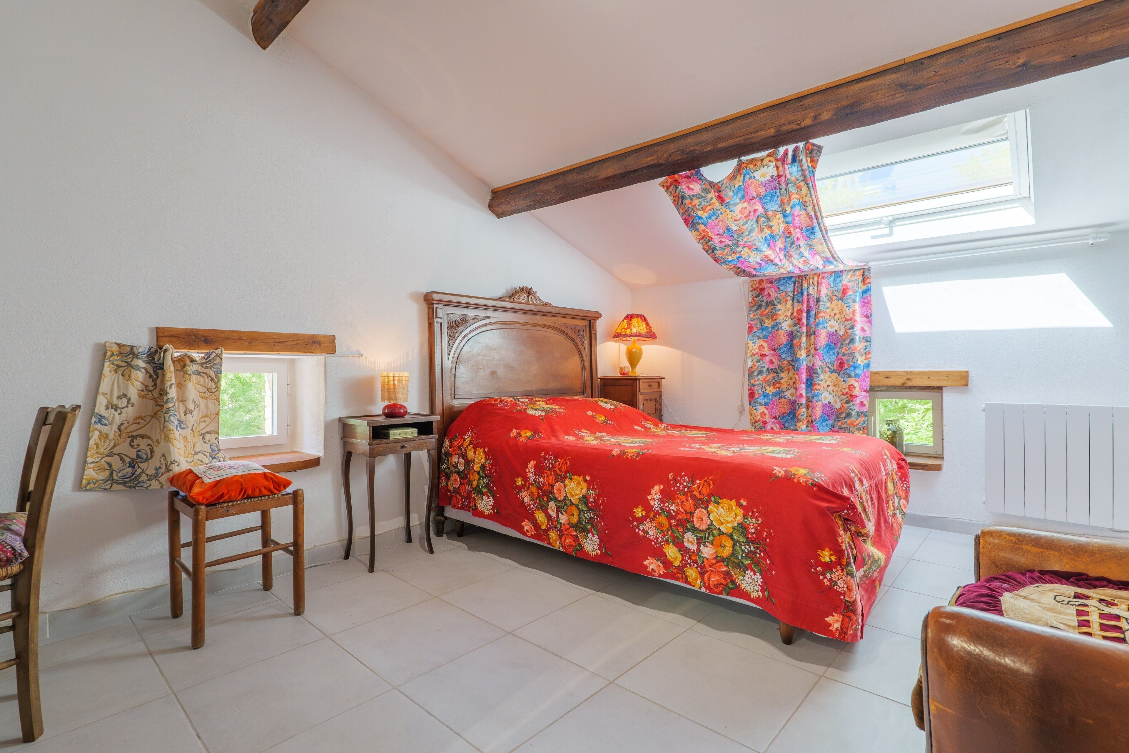 A bedroom with a bed, a chair, a nightstand, a lamp, and a window with a curtain.