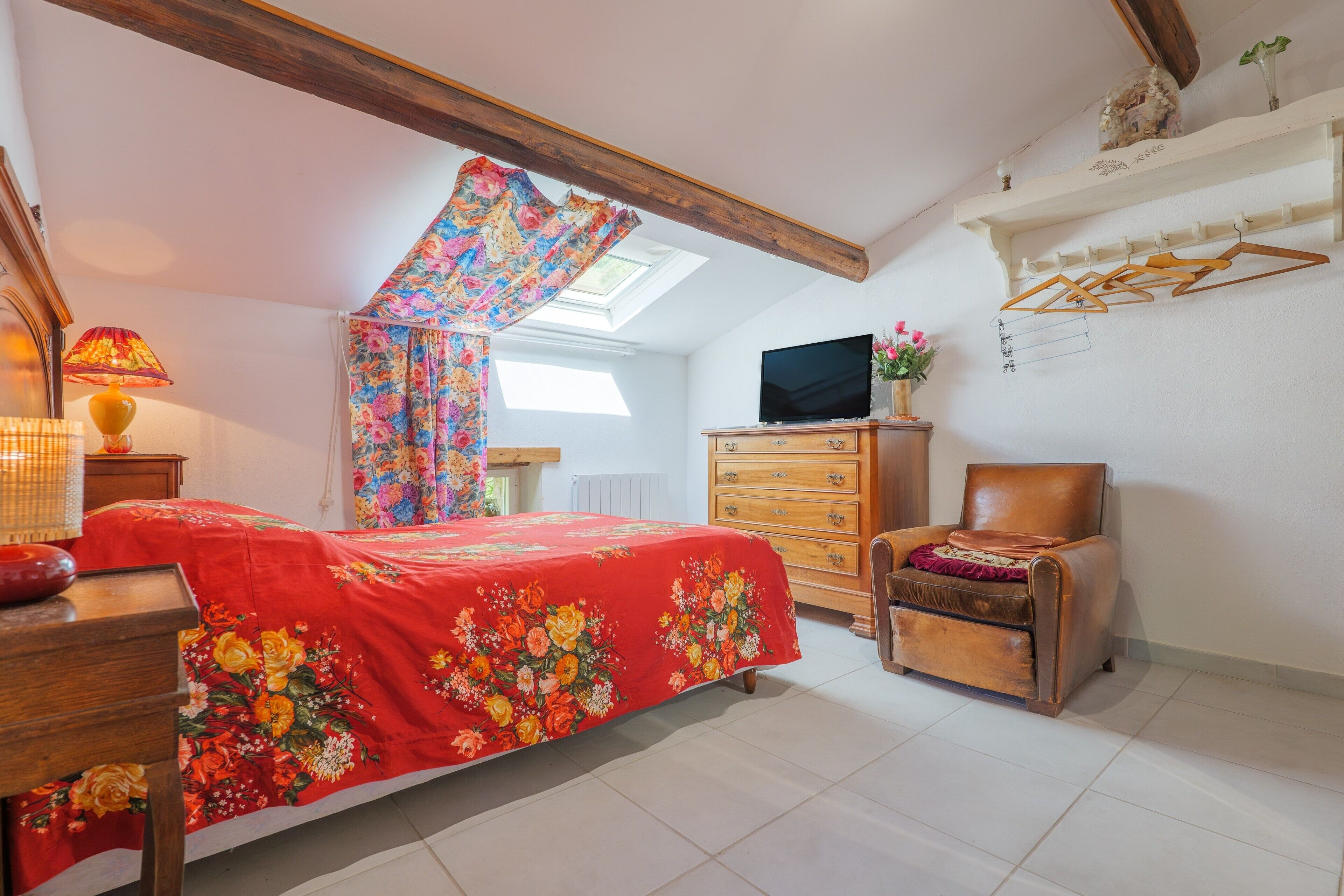 A bedroom with a bed, a wooden dresser, a leather armchair, a television, and a skylight.