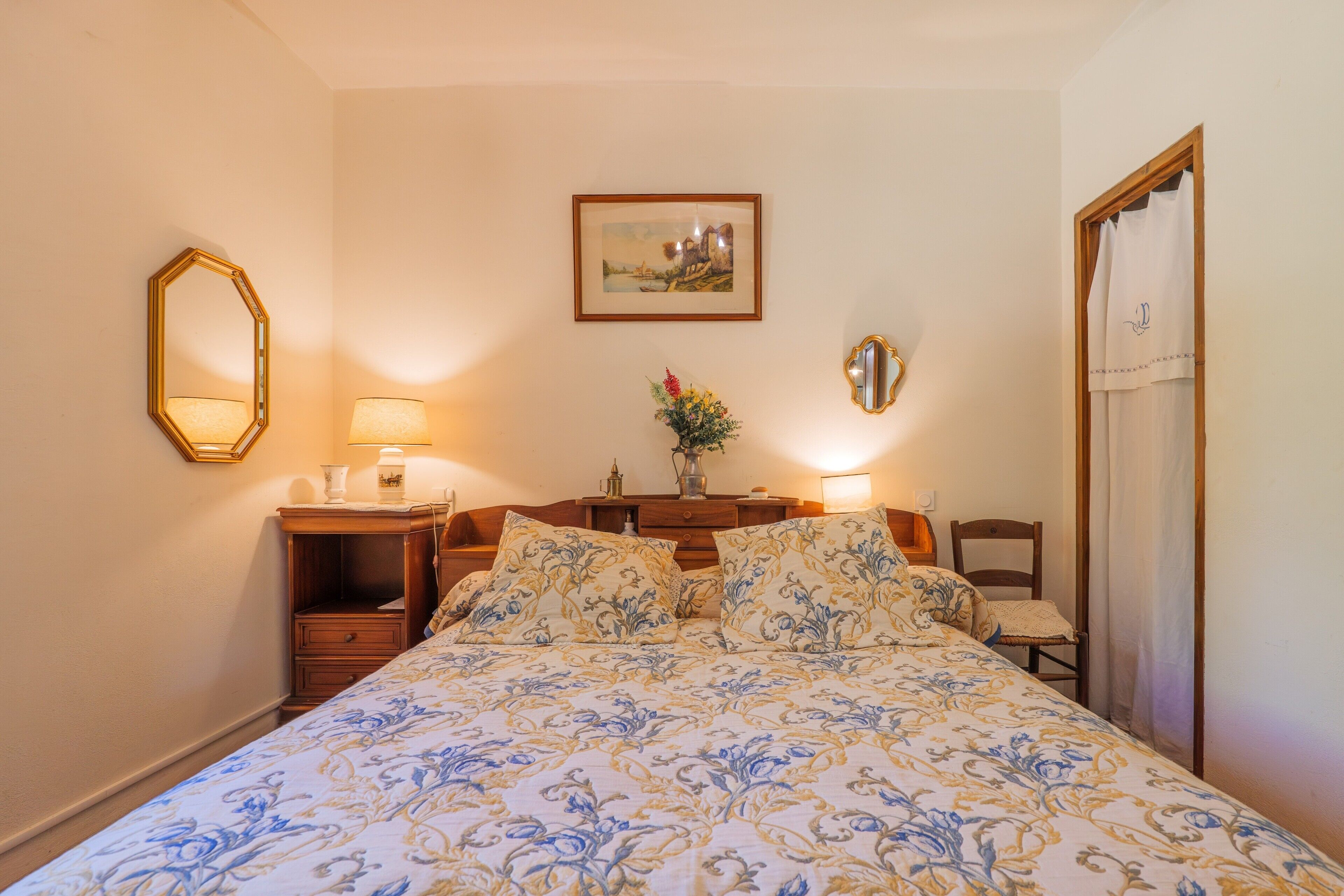 Ein Schlafzimmer mit Bett, Nachttischen, Lampen, einem Spiegel, einem Bilderrahmen und einer Tür.