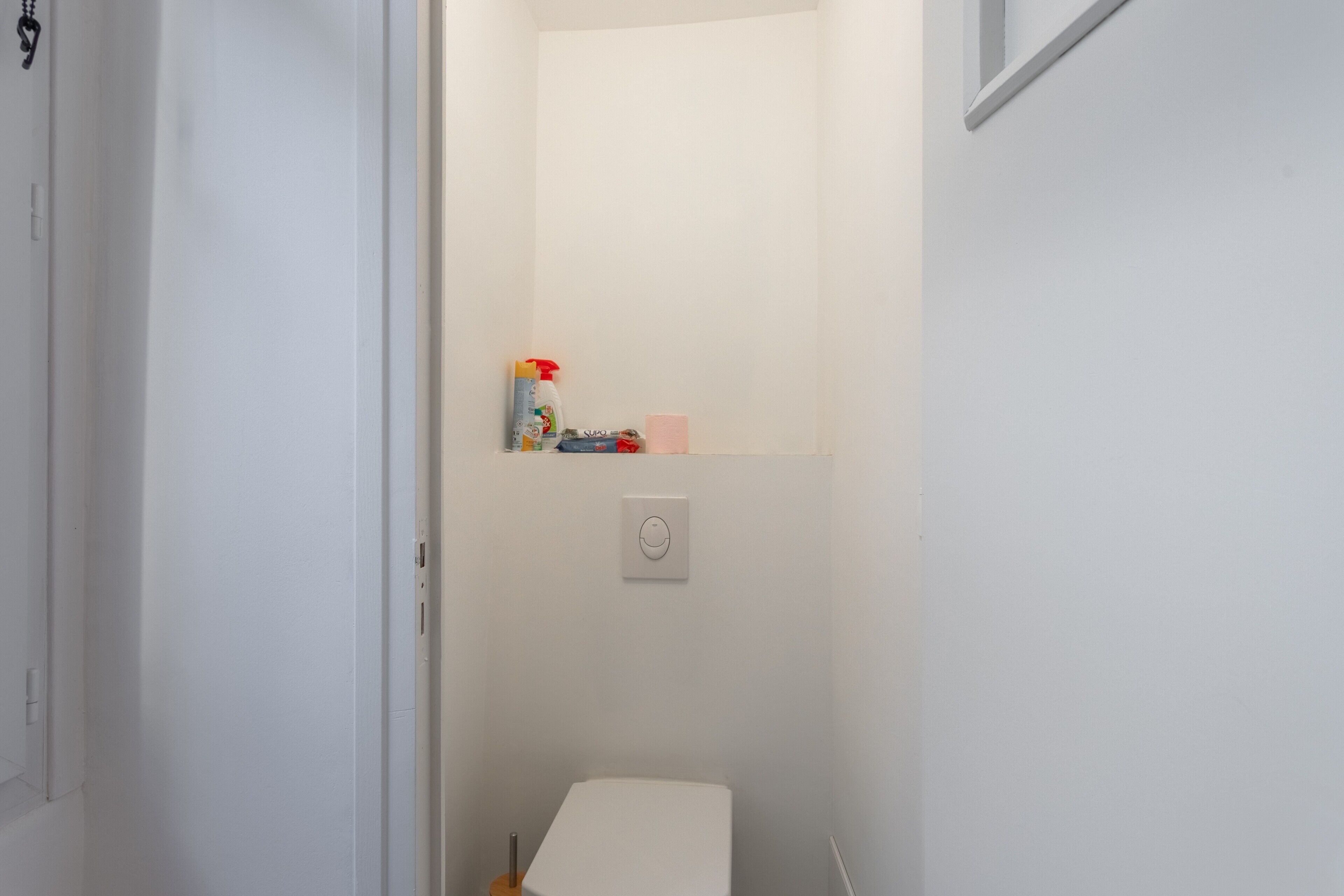 Un baño pequeño y blanco con inodoro, un estante con artículos de tocador y un interruptor de luz.
