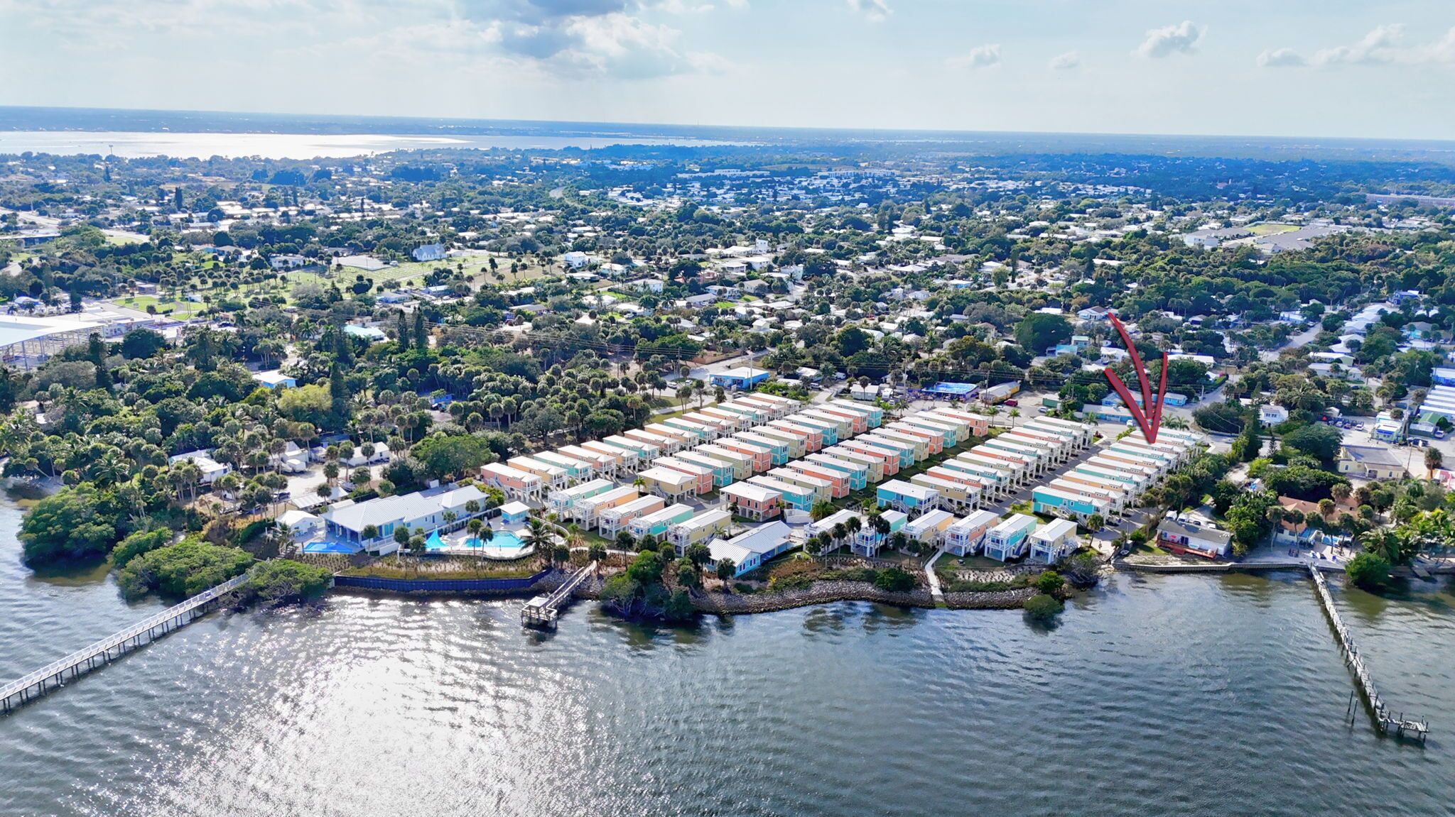 An aerial view of a coastal area with a row of colorful mobile homes by the water, a bridge, and a distant cityscape.