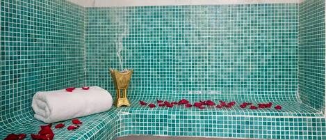 A corner bathtub with teal mosaic tiles, a white towel, a golden decorative item, and scattered rose petals.