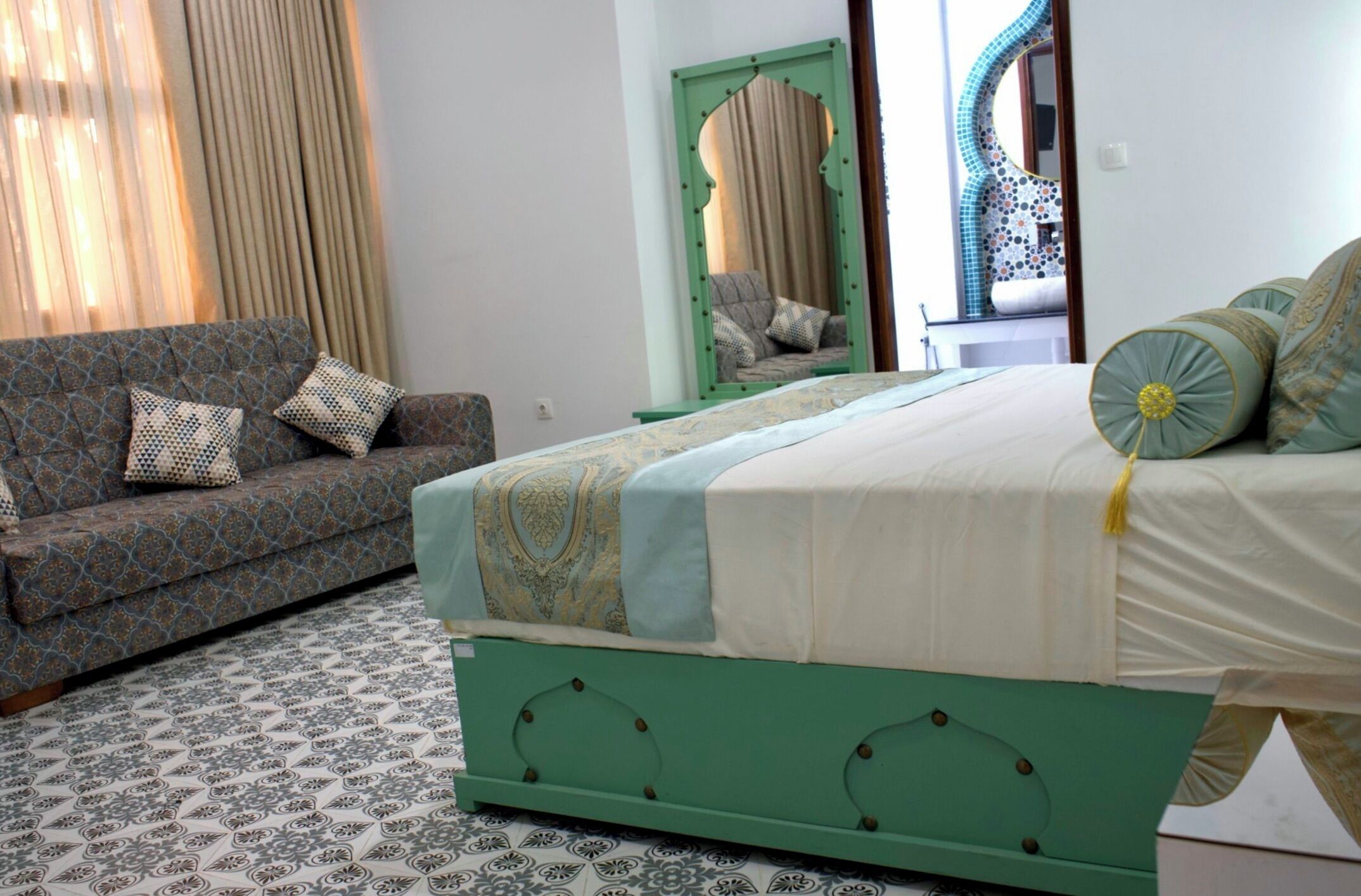Un dormitorio con piso estampado, una cama verde con colcha blanca y amarilla, un sofá estampado con almohadas, un espejo y una ventana con cortinas.