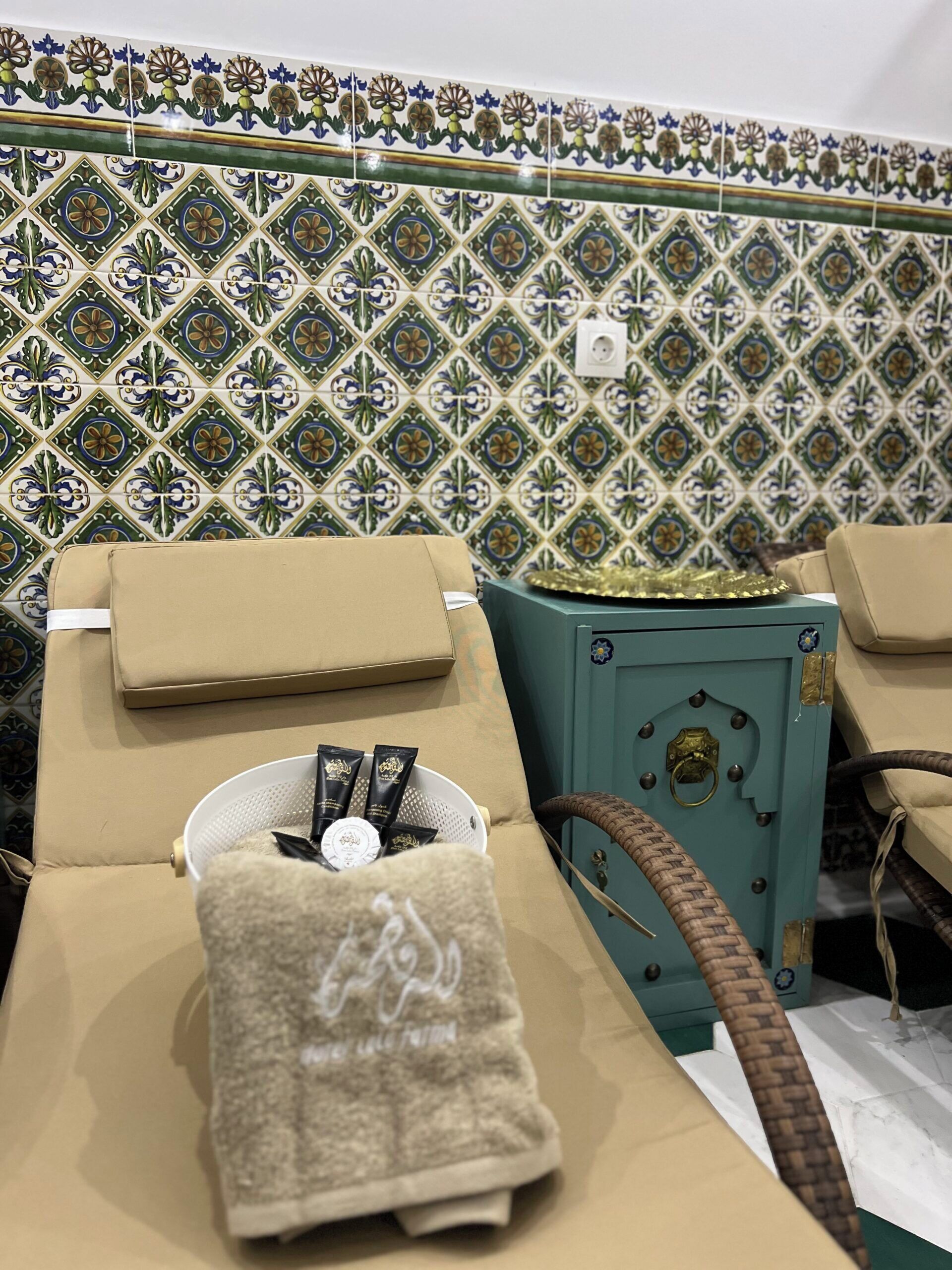 Ein Zimmer mit Wand aus gemusterten Fliesen, einem Massagetisch mit Handtuch, einem kleinen Korb mit Gegenständen und einem türkisblauen Schrank.