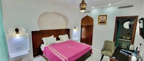 A bedroom with a bed, a chair, a desk, a mirror, and a decorative ceiling light.