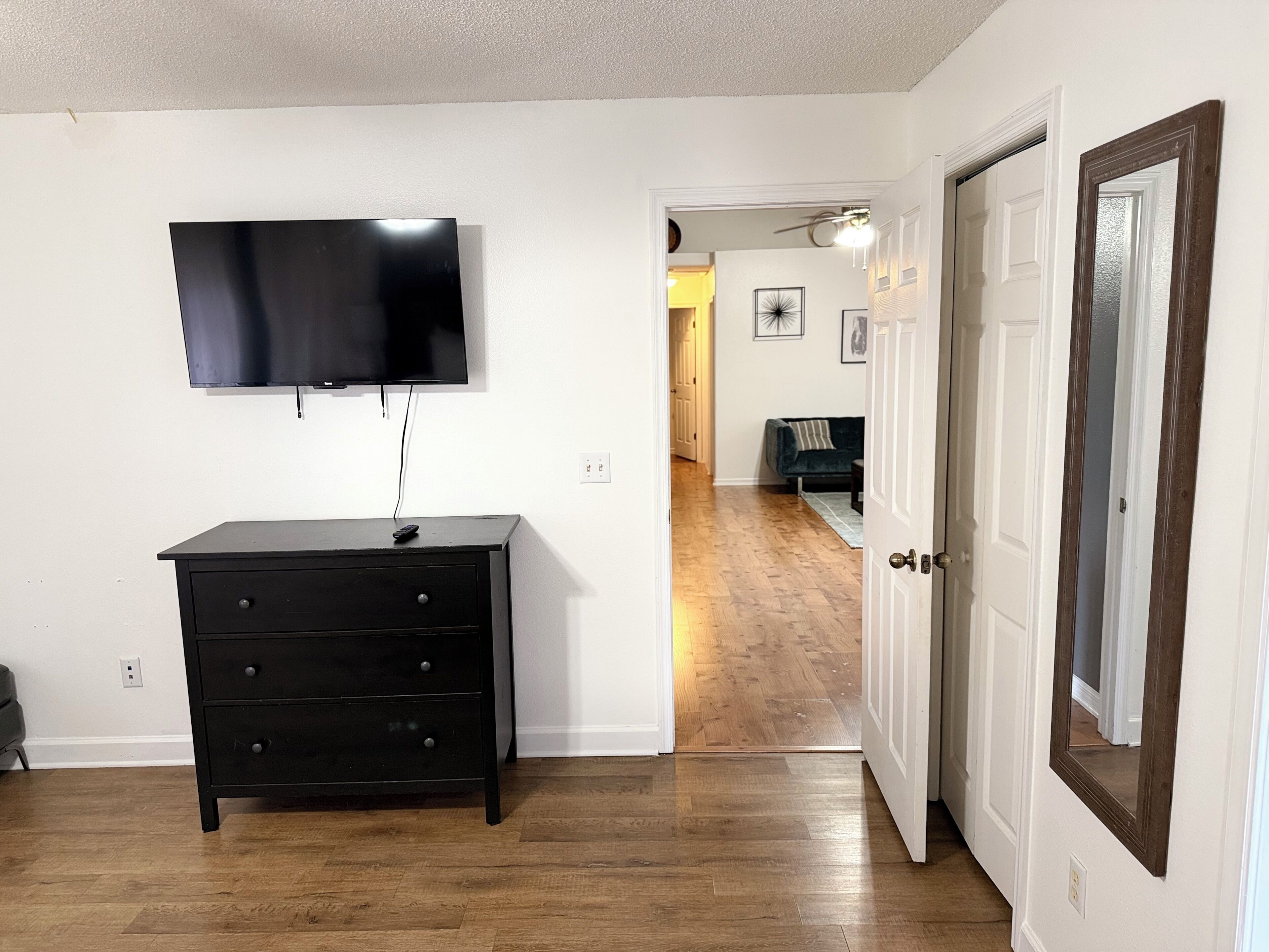 A room with a flat-screen TV mounted on the wall, a black dresser with drawers, a wooden floor, and a white door with a mirror.