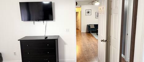 A room with a flat-screen TV mounted on the wall, a black dresser with drawers, a wooden floor, and a white door with a mirror.