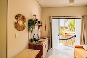 A modern kitchen with a built-in island, a dining area, and a sliding glass door leading to a balcony with a hammock. - Studio Graviola with ocean view / prime location / parking / rooftop (Itacaré)