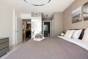 Ein modernes Schlafzimmer mit einem großen Bett, einer Kommode, einem durchgehenden Türbogen sichtbaren Badezimmer und einem Essbereich im Hintergrund.