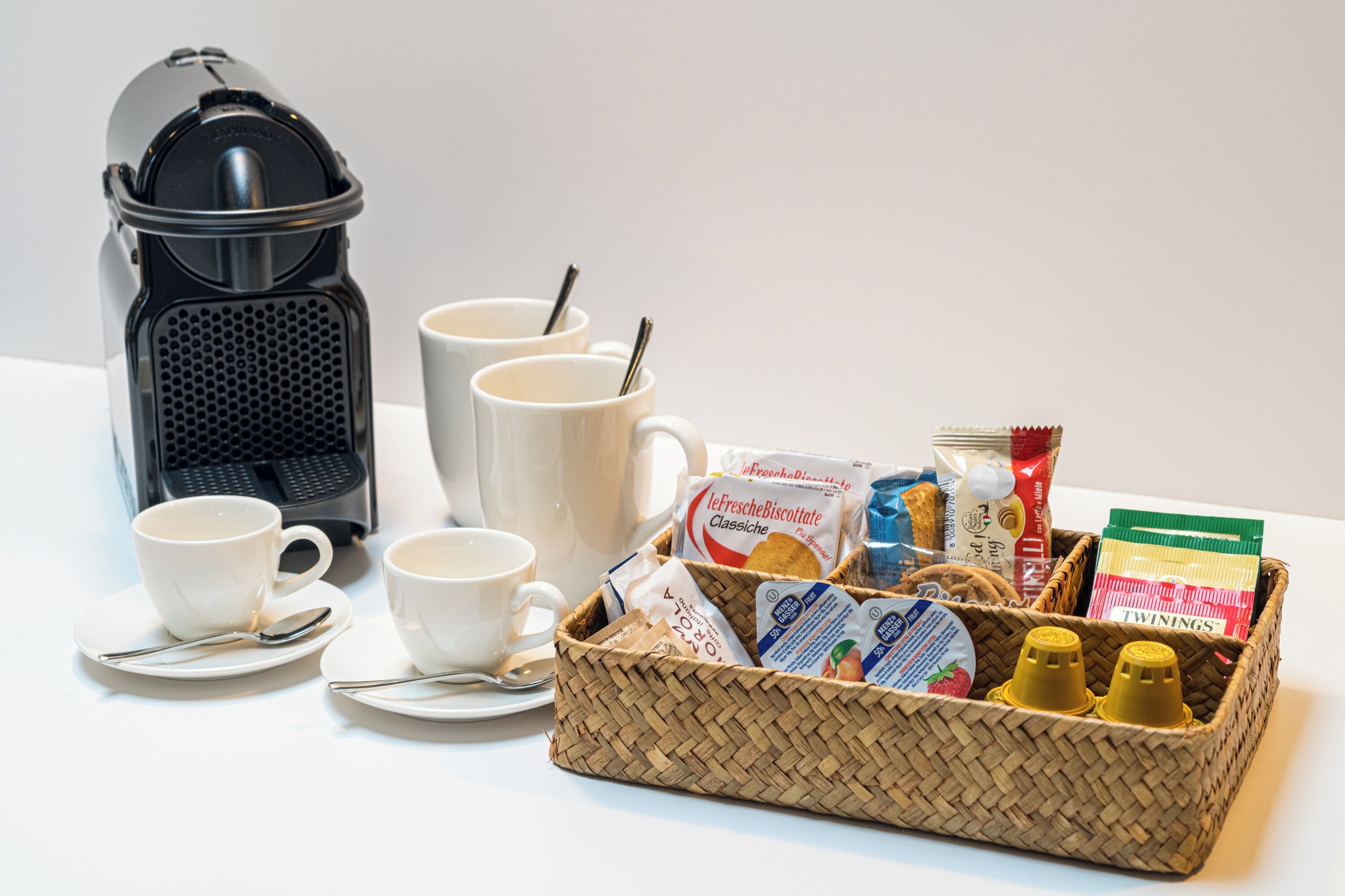 A breakfast setup with a coffee maker, cups, a basket of food items, and condiments.