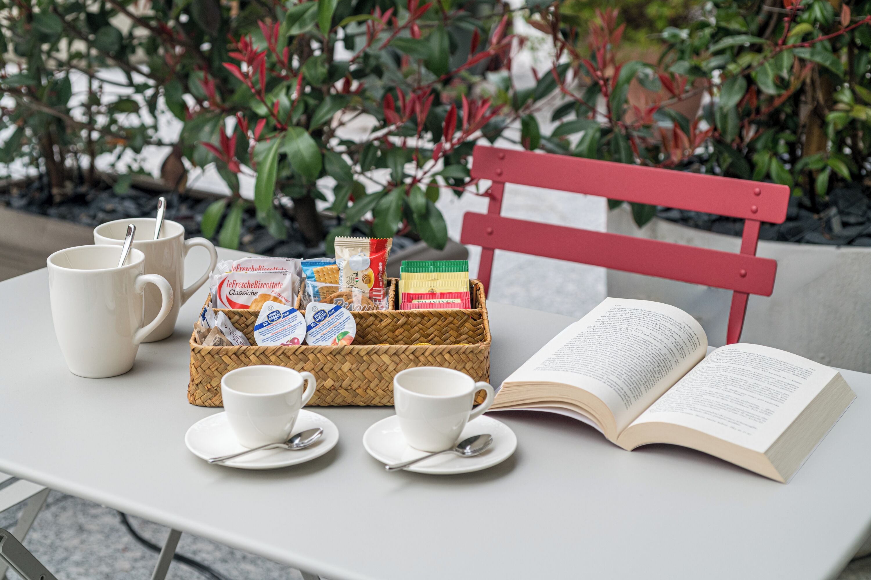 A table with a basket of snacks, two cups with saucers, a spoon, and an open book.