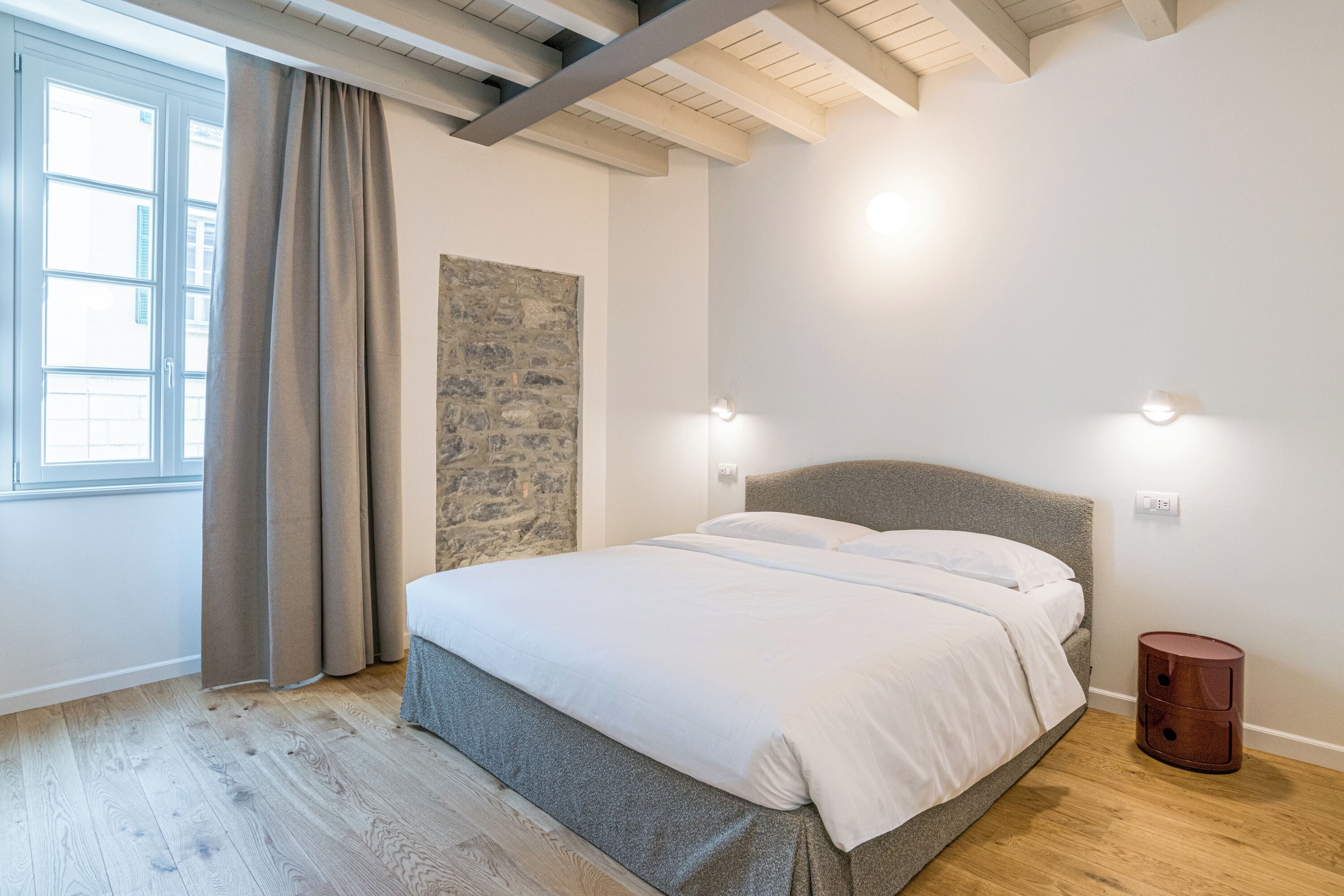 Une chambre avec un lit, un mur en pierre servant de décoration, une fenêtre avec des rideaux et un plancher en bois.