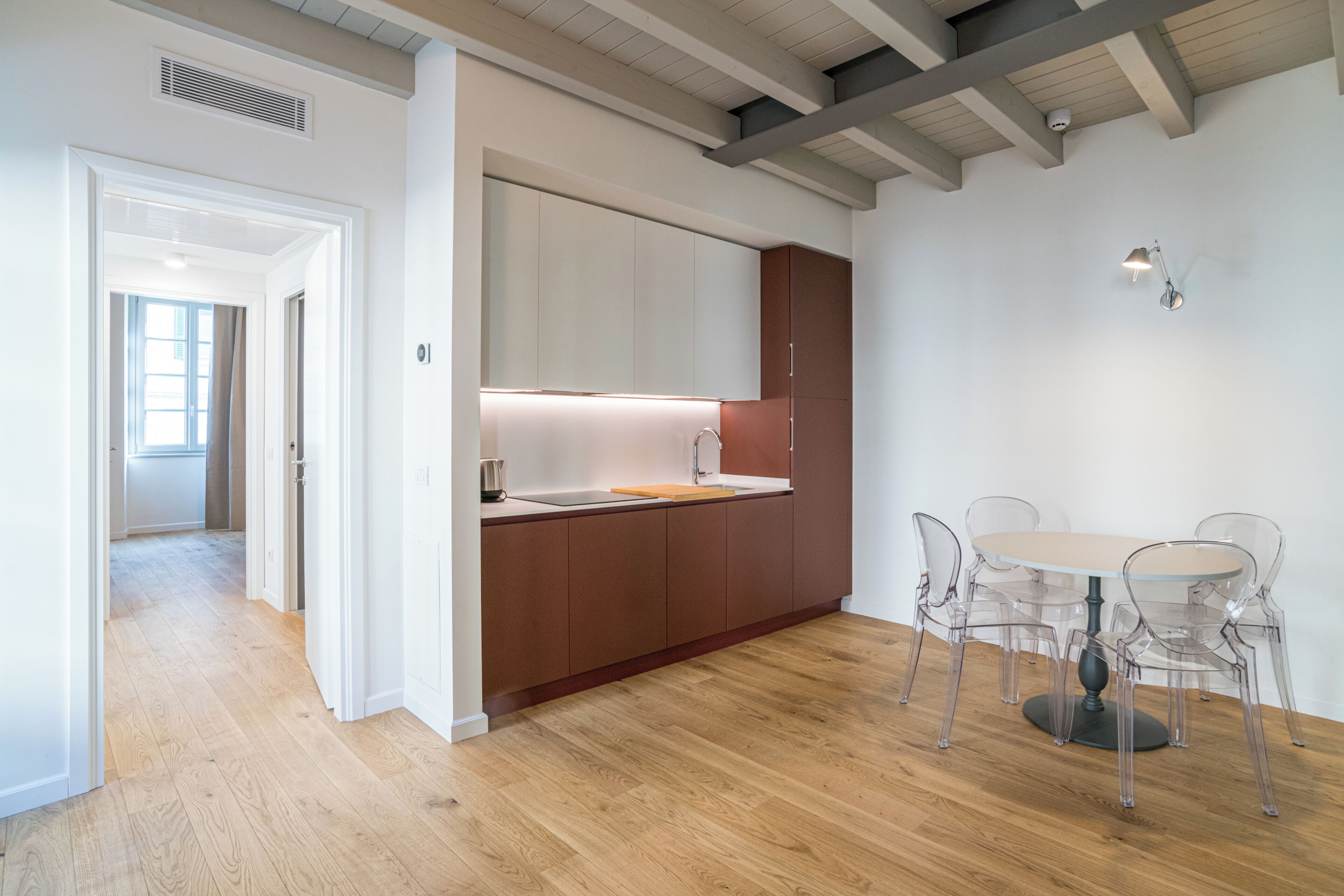 A modern kitchen with wooden flooring, a dining area with clear chairs, and a white ceiling with exposed beams.