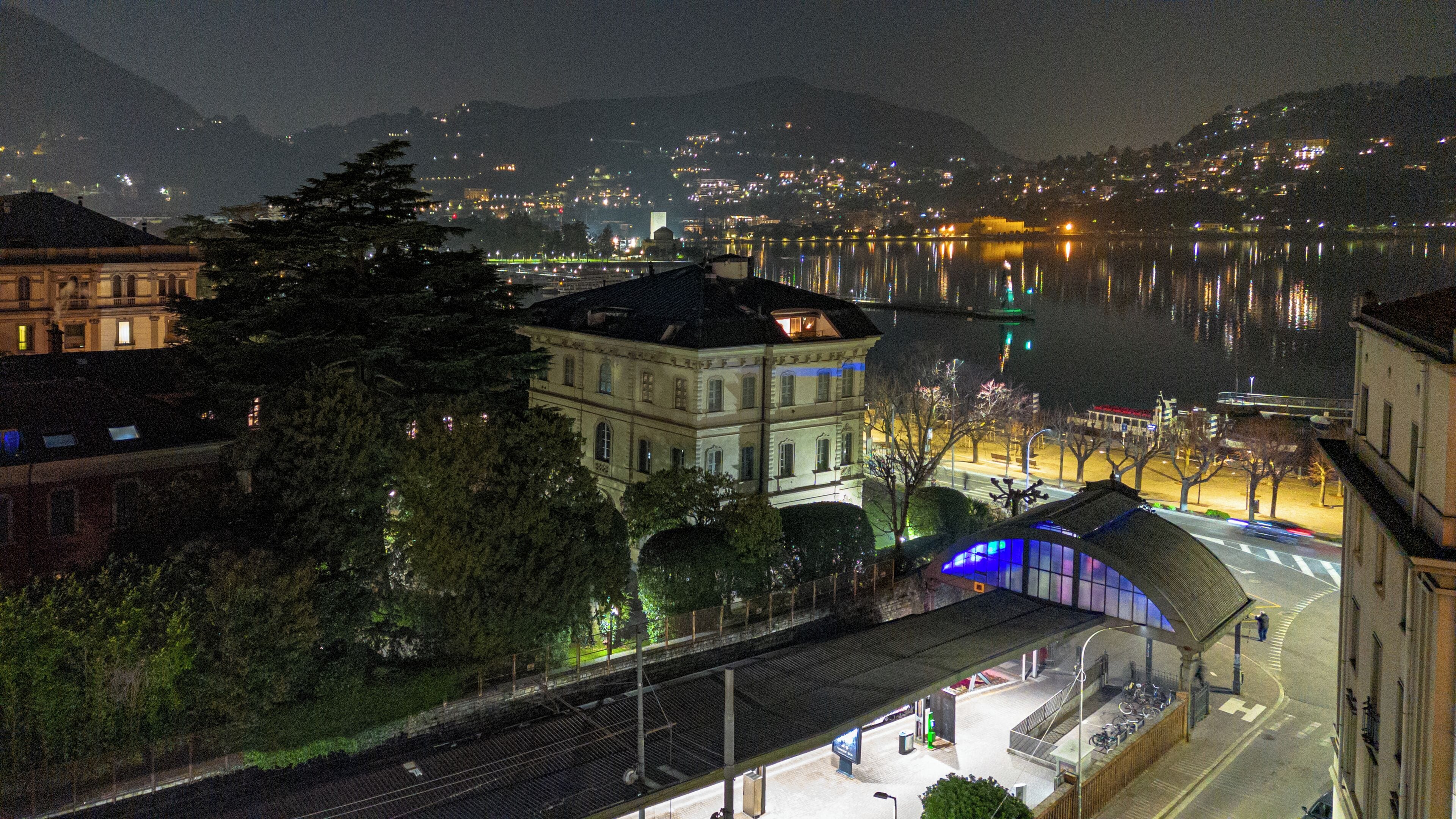 A nighttime view of a waterfront cityscape with buildings, a train station, and a body of water reflecting lights.