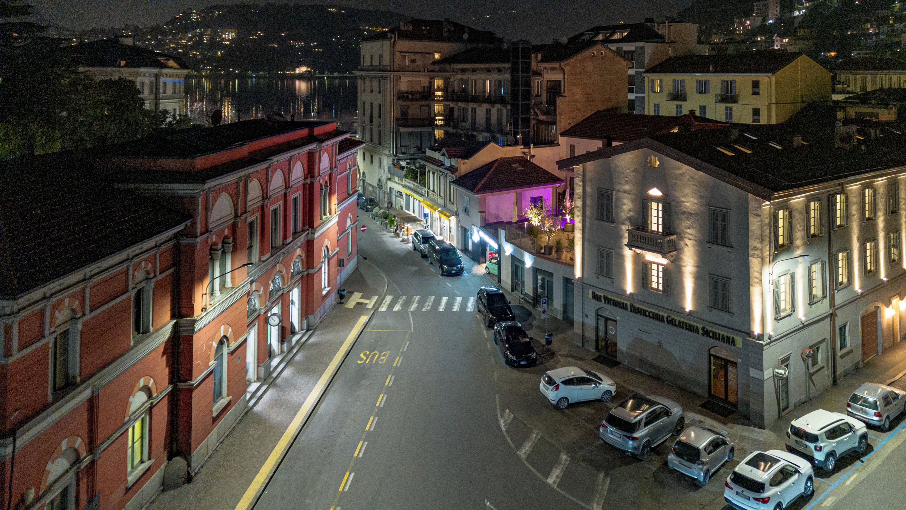 Une vue nocturne d’une rue bordée de voitures garées et d’immeubles dont les enseignes sont illuminées.