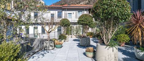 A courtyard with potted plants, a stone floor, and a building with a red roof in the background.
