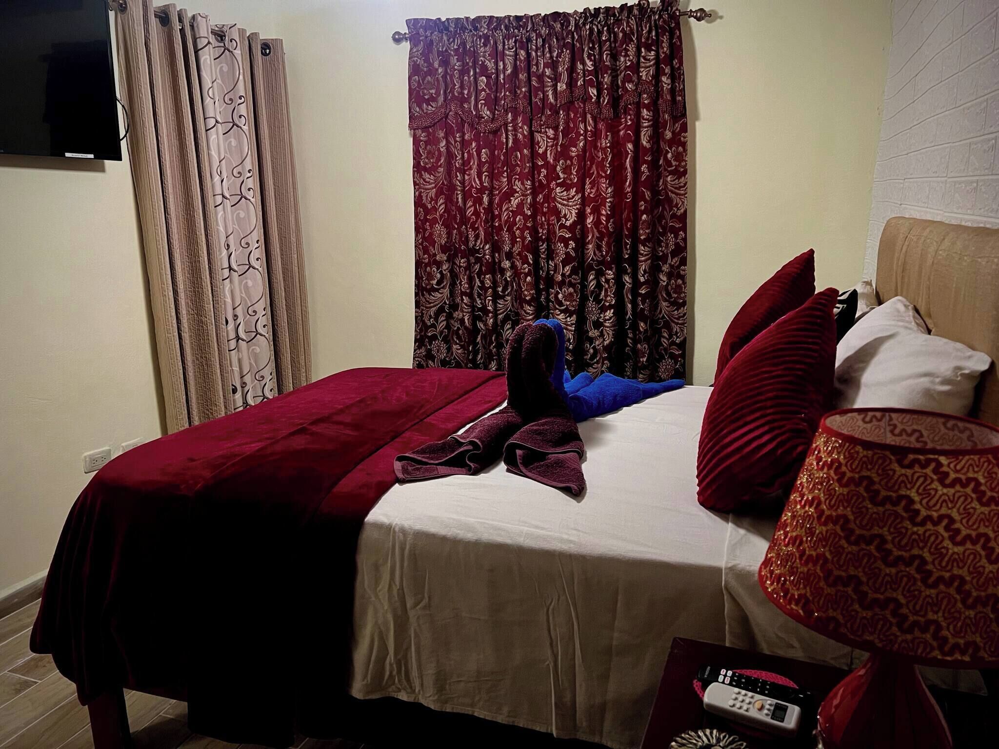 A bedroom with a bed, pillows, a blanket, a remote control, and curtains.