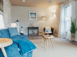A cozy living room with a blue sofa, a small wooden table, a chair, a lamp, and a window with curtains. - 4 Star Holiday Home in Sagad (Sagard)