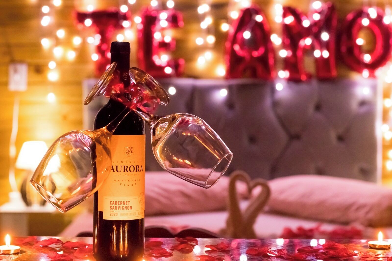 Una botella de vino con copas, sobre una mesa con pétalos de rosa y velas, con una cama y decoraciones que dicen 'TE AMO' al fondo.