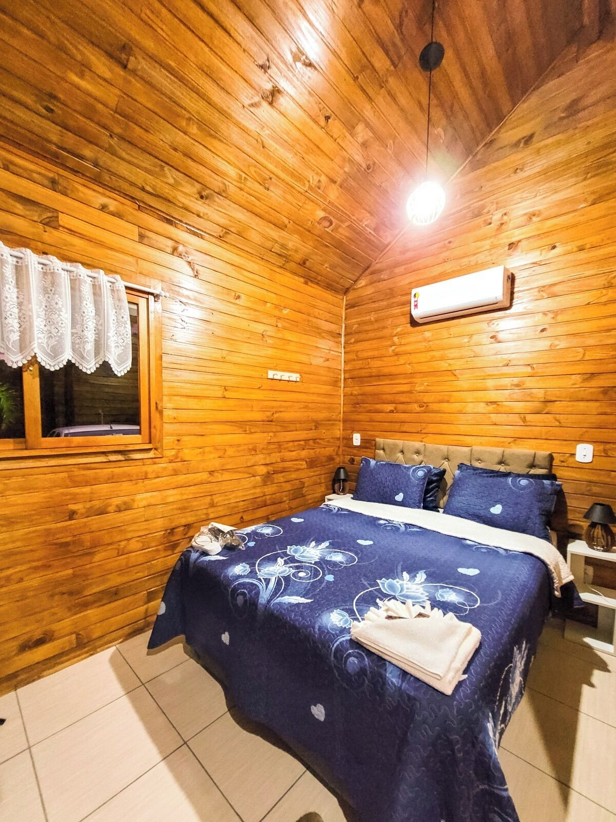 Une pièce aux murs lambrissés, avec un lit, une fenêtre ornée d’un rideau en dentelle et un appareil de climatisation.