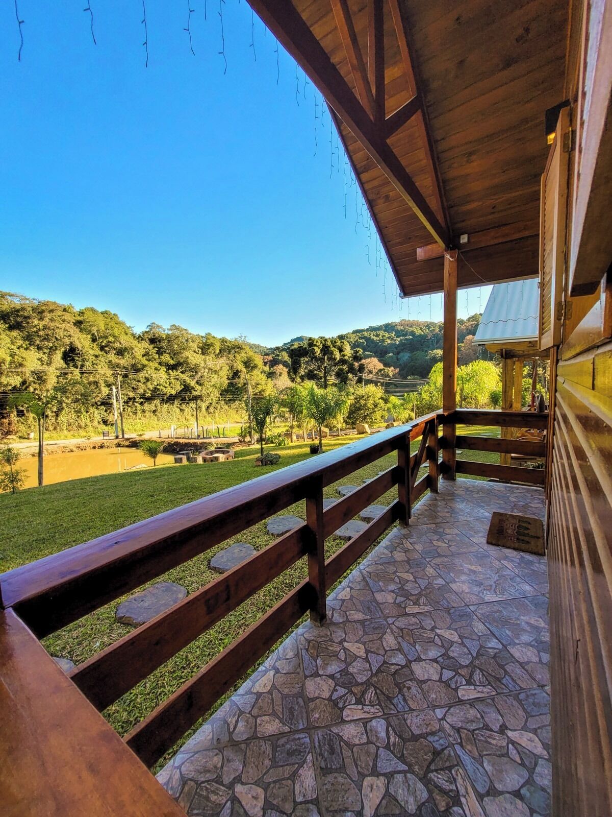 A covered outdoor area with a stone floor, wooden railing, and a view of a green landscape with trees and hills.