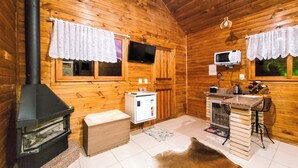 Interior de una cabaña de troncos con estufa de leña, kitchenette y un ventanal grande.
