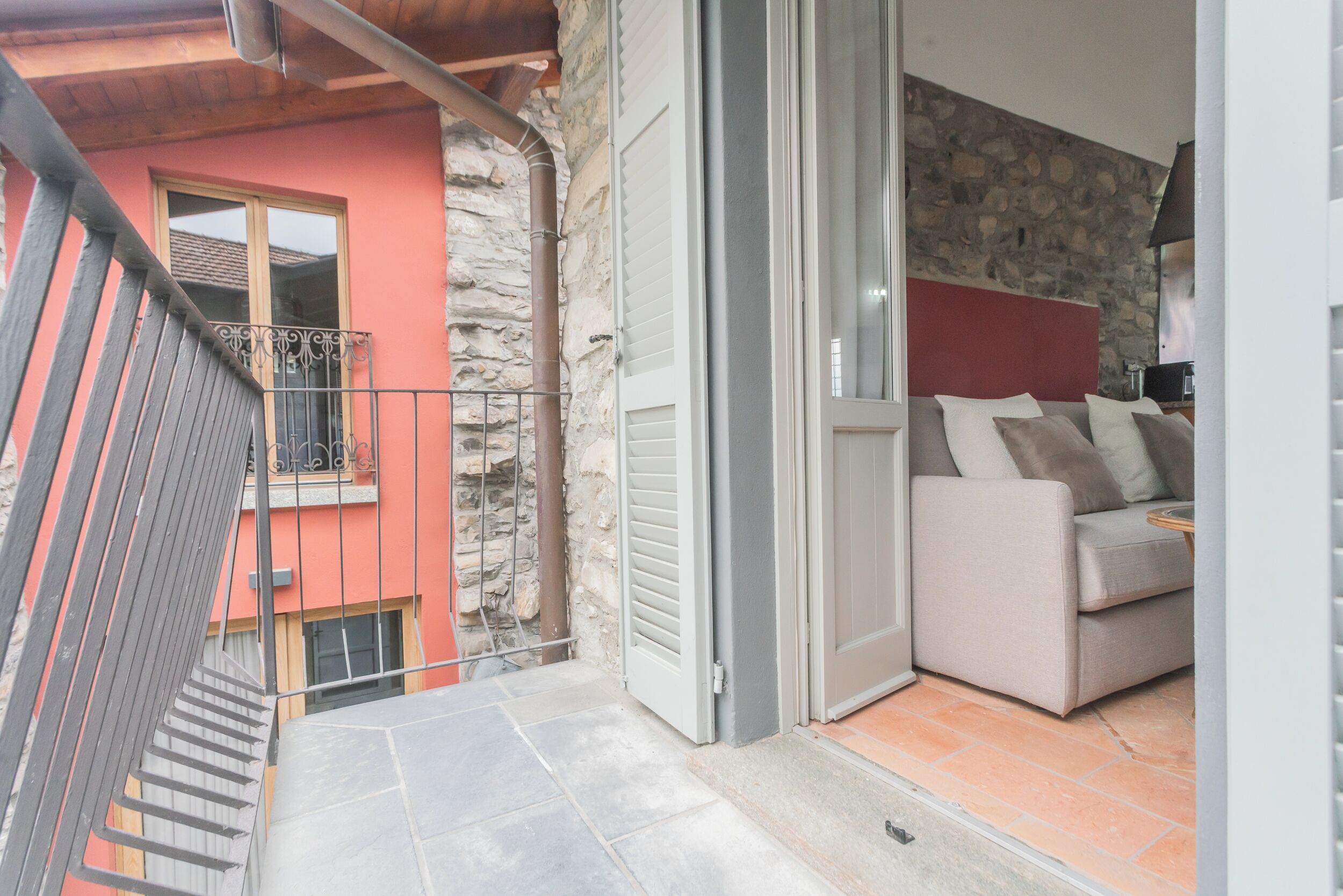 Un balcone con ringhiera in metallo, un muro in pietra, un soffitto in legno e un divano beige con cuscini.