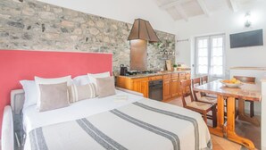 A bedroom with a stone wall, a red headboard, a bed with multiple pillows, a wooden dining table, and a kitchen area with wooden cabinets and a stove.