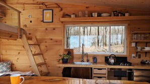 A wooden cabin interior with a kitchen, a window with a view of trees, a ladder, and a bed with a pillow. - Penner Cabin | Peaceful Wilderness Retreat (Minnow Drive, Dysart et al, ON)