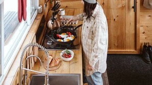 A kitchen with wooden cabinets, a stove, and a person cooking. - Penner Cabin | Peaceful Wilderness Retreat (Minnow Drive, Dysart et al, ON)