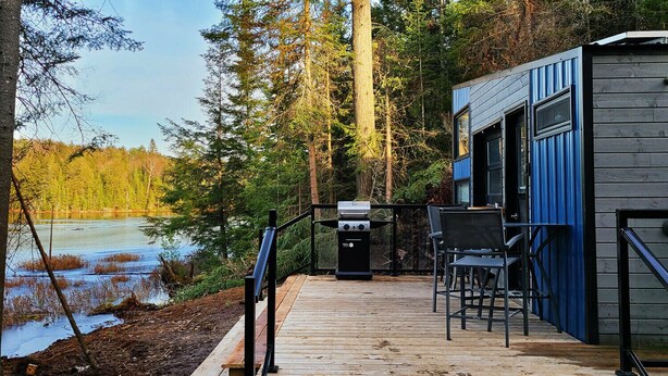 A deck with a grill, chairs, and a view of a forested area and lake. - Penner Cabin | Peaceful Wilderness Retreat (Minnow Drive, Dysart et al, ON)
