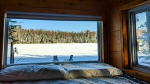 A wooden cabin room with a bed, large windows, and a view of a snowy landscape. - Penner Cabin | Peaceful Wilderness Retreat (Minnow Drive, Dysart et al, ON)