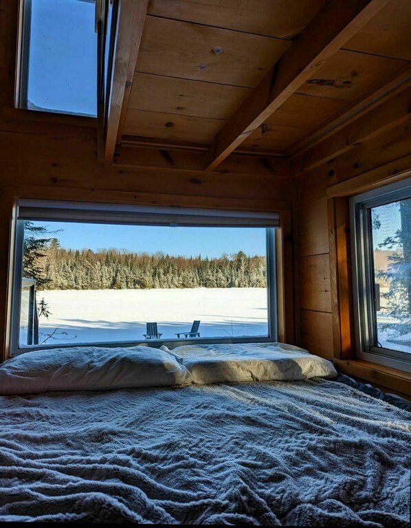 A wooden cabin room with a bed, large windows, and a view of a snowy landscape.