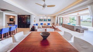 A spacious living area with a large wooden dining table, white sofas, and a view of a garden through large windows. - Ground Floor Punta Mita Condo with Shared Pool (Punta Mita)