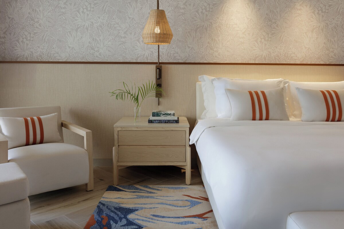 A hotel room with a bed, a chair, a nightstand with a plant and book, and a patterned wall.