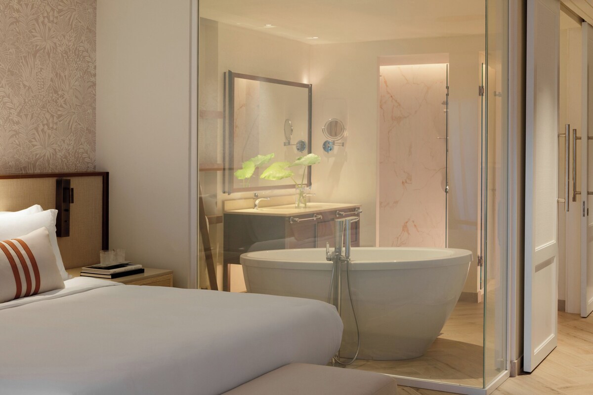 Een moderne hotelkamer met een groot bed, een badkamer met glazen wanden en een vrijstaand bad, een wastafel en een spiegel.