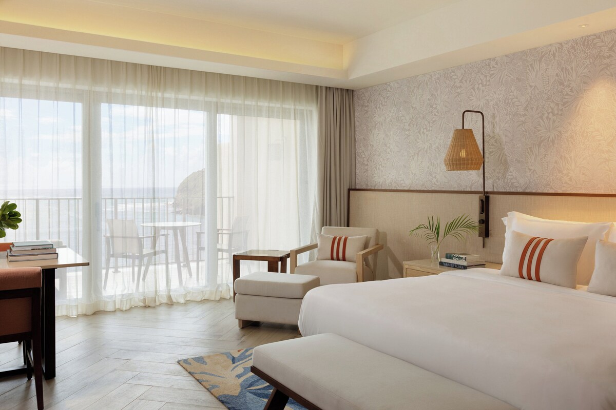 A hotel room with a large bed, a desk with books, a chair, a small table, and a view through sheer curtains.