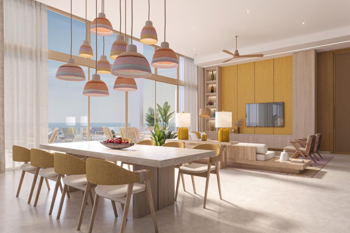 A modern dining area with a large table, multiple chairs, pendant lights, and a view of the ocean.