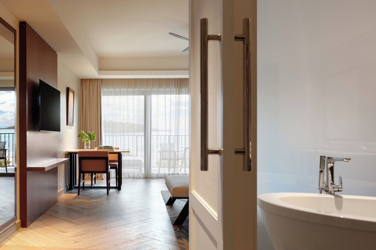 A modern hotel room with a flat-screen TV, a dining area with a table and chairs, a large window with curtains, and a bathroom with a bathtub.