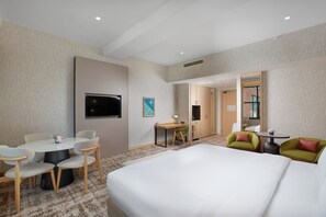 Quarto de hotel com cama, escrivaninha, duas cadeiras, televisão e um espelho grande.