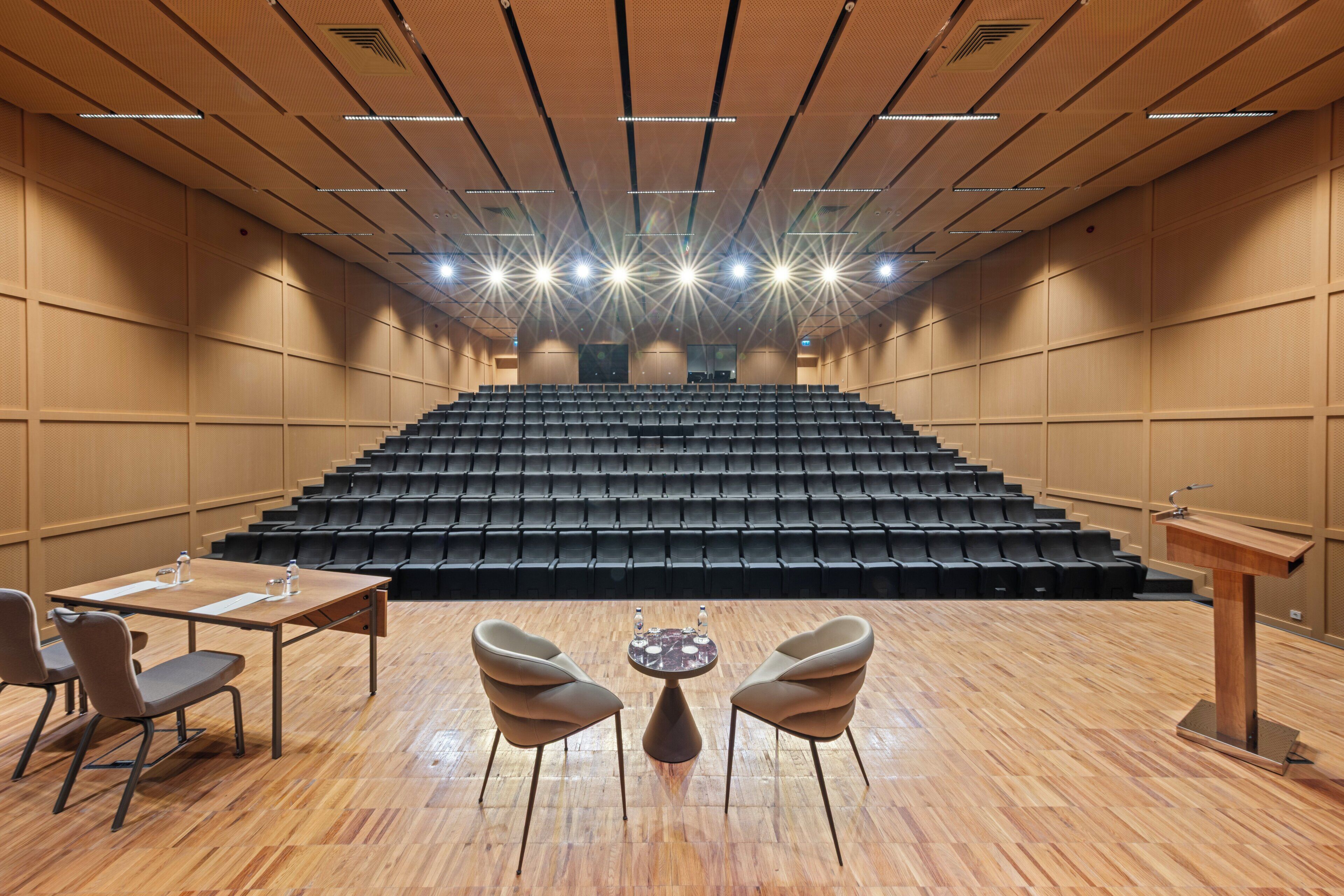 Un salón de conferencias con un escenario, filas de sillas, un atril y una mesa con sillas.