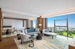 Ein großzügiges Wohnzimmer mit einer Couch, Sesseln, einem Couchtisch und einem Essbereich. Große Fenster bieten Blick auf die Stadt.