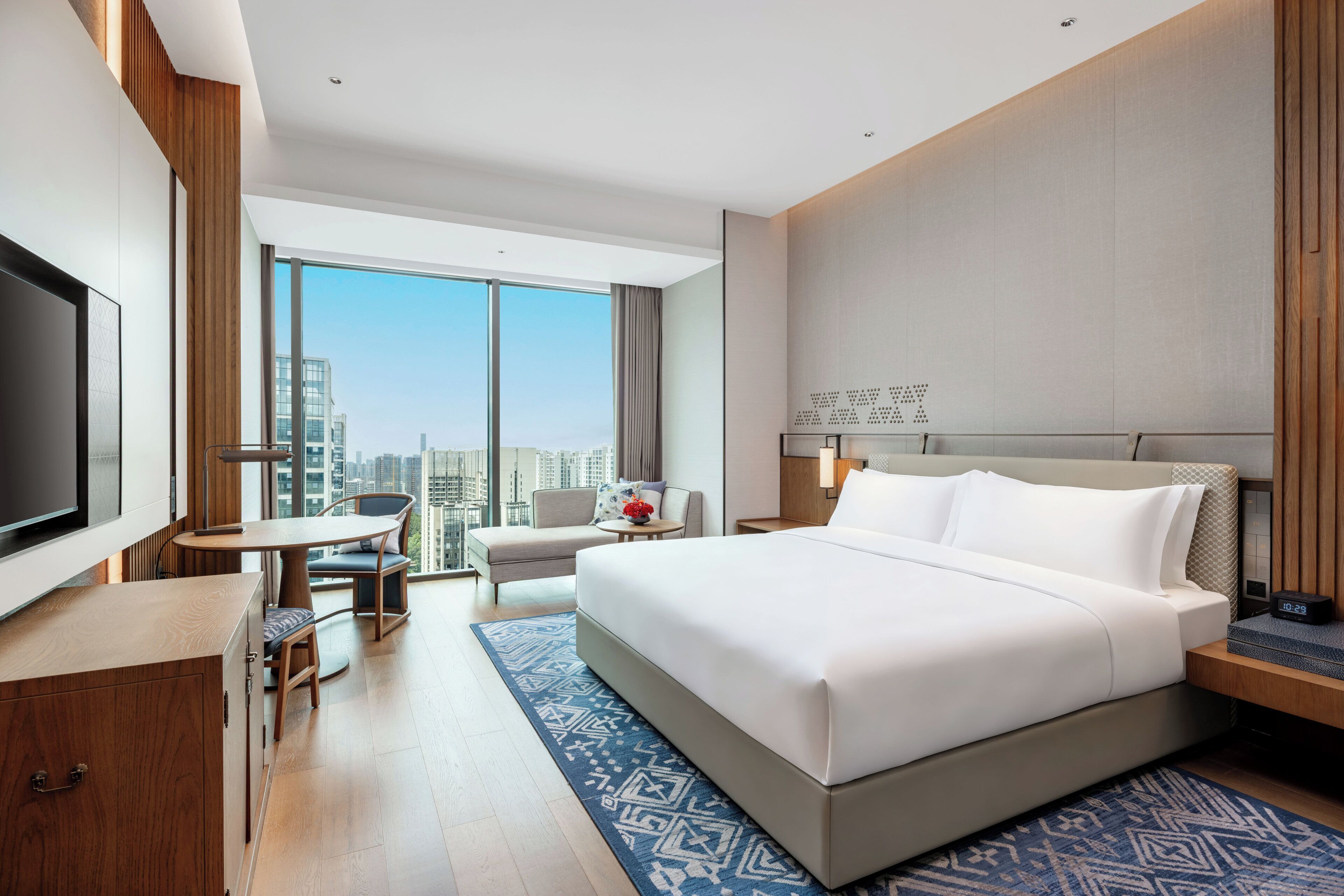 A modern hotel room with a large bed, a desk, a chair, a TV, and a view of the city.