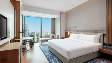 A modern hotel room with a large bed, a desk, a chair, a TV, and a view of the city.