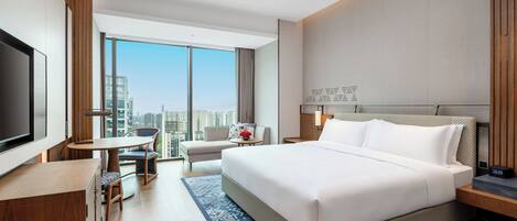 A modern hotel room with a large bed, a desk, a chair, a TV, and a view of the city.