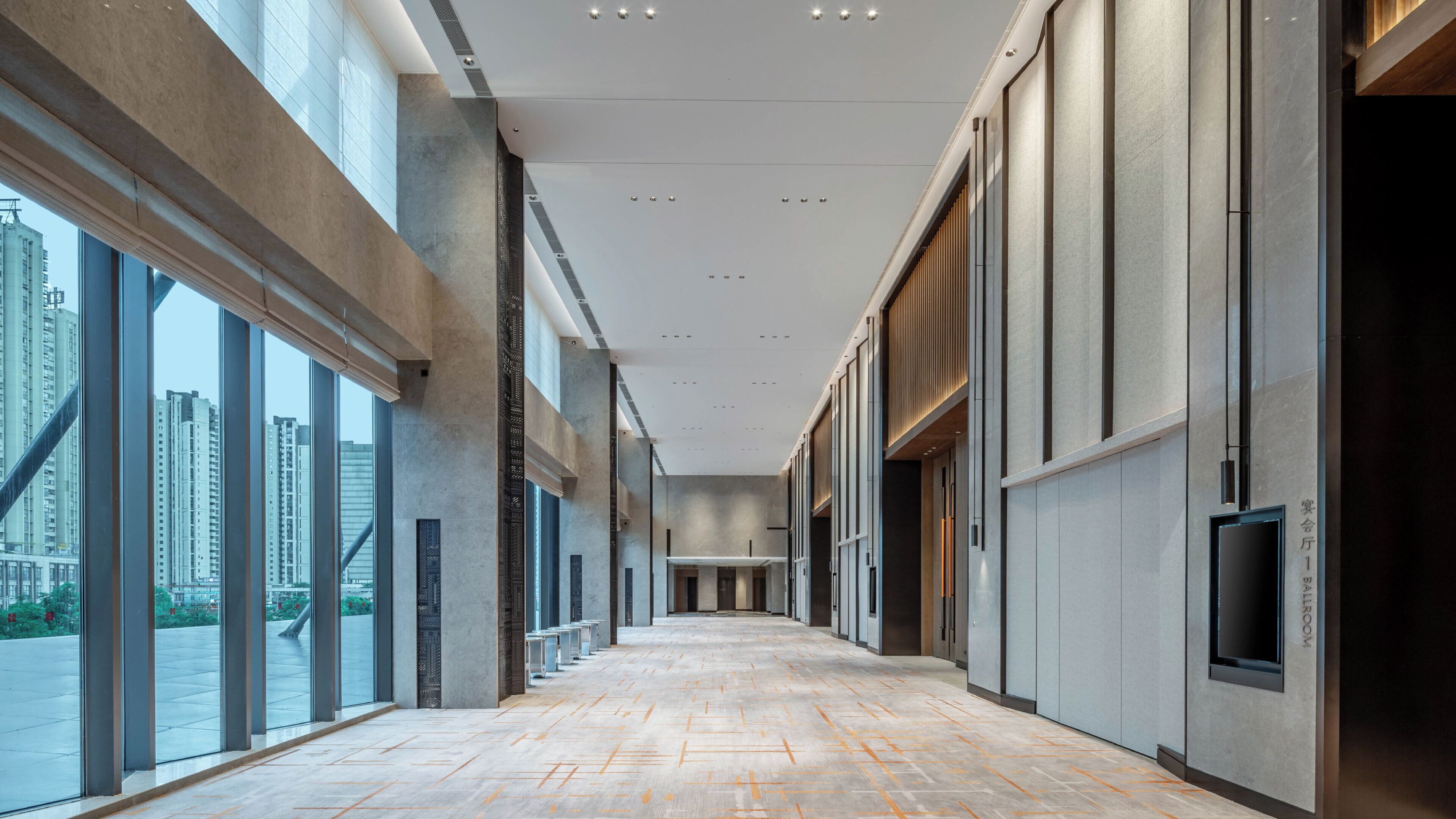 A long, narrow hallway with large windows on one side and a wall-mounted television on the other.
